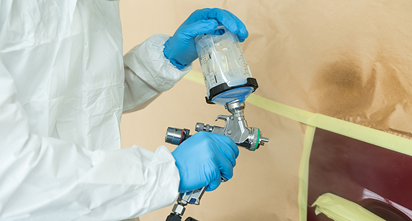 Person wearing a protective suit and blue gloves preparing a spray gun for automotive painting. The car panel is masked off with paper and tape, ready for paint application.